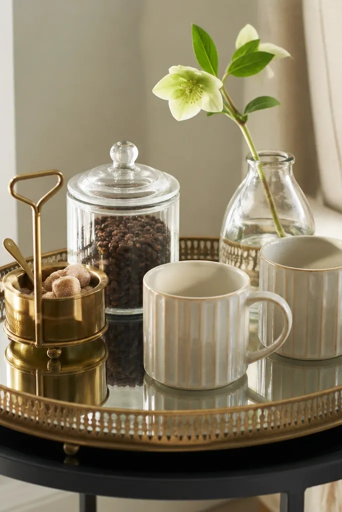 A side table holds a large mirrored tray. On the tray is a gold-toned caddy with sugar, a glass jar of beans, a small vase with one stem, and two matching mugs. The mirrored surface reflects the items and light.