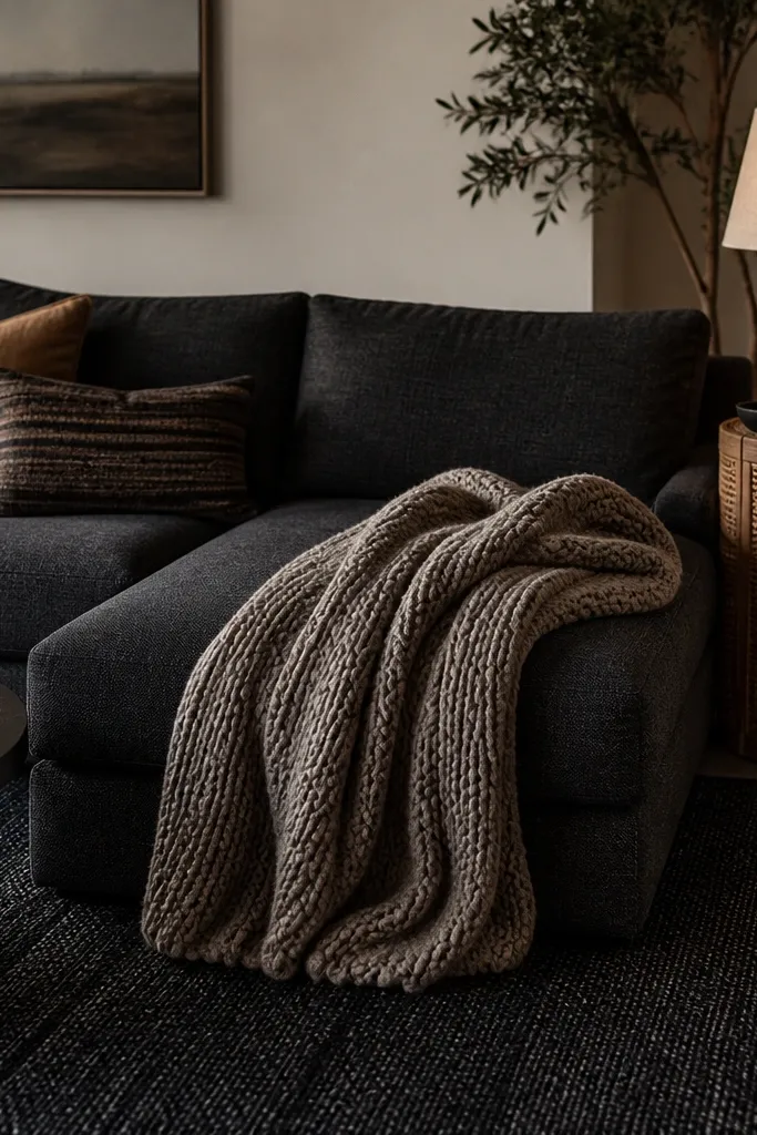 A dark charcoal sofa with a thick knit throw draped over the chaise. A rattan side table sits next to the sofa, and there's a warm amber glass lamp on top. The rug is dark with a subtle woven pattern.