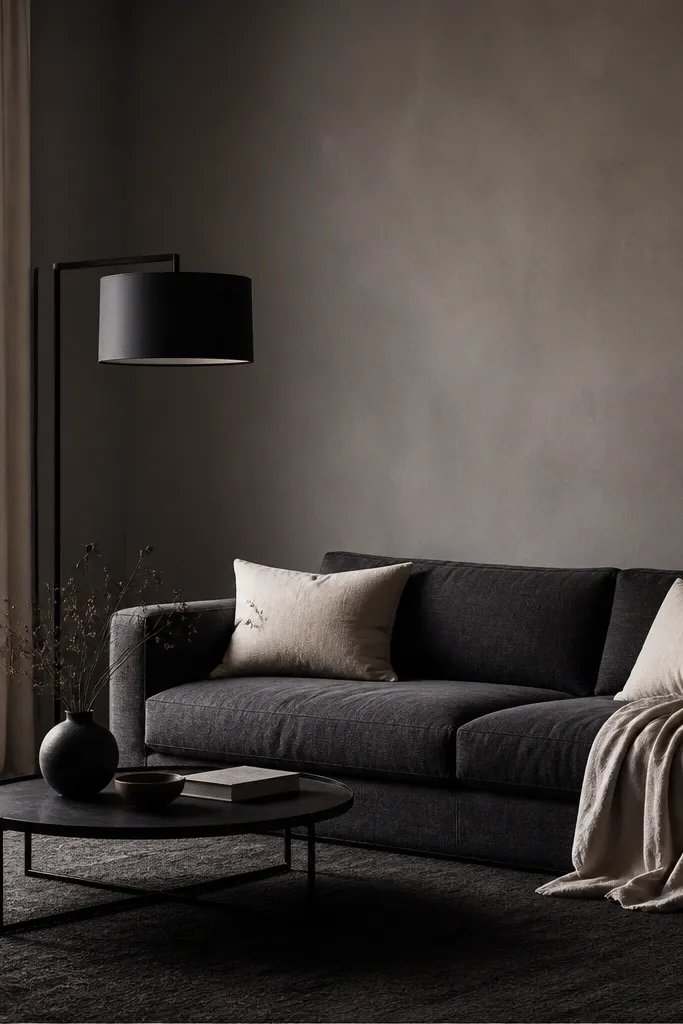 Smoky grey walls with a dark grey sofa. A black metal floor lamp with a drum shade stands beside the seating. The rug is charcoal, and cushions are cream with a soft linen texture.