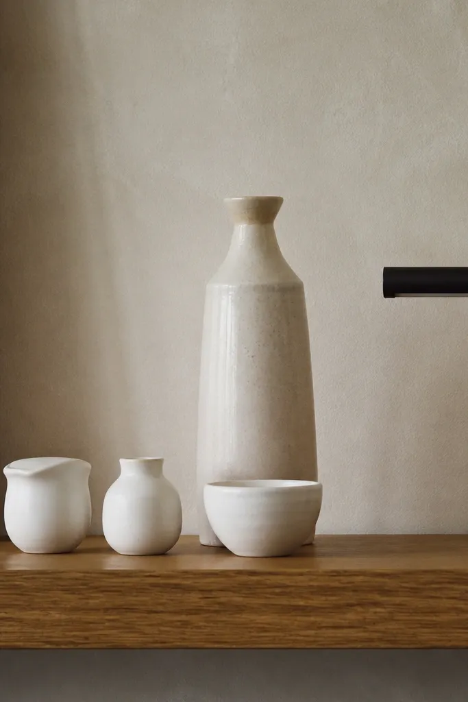 Three small ceramic objects in matte white line up along the shelf front, with a taller off-white ceramic vessel behind them. A thin black metal picture light sits at the far right.