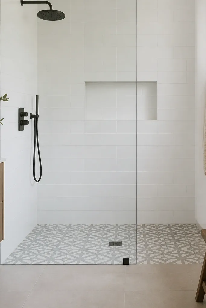 A walk-in shower with simple white wall tile and a patterned floor tile with a geometric motif inside the shower pan only. The niche is a plain rectangular cutout with white tile. The glass is frameless, and the rest of the bathroom floor outside the shower is plain neutral tile.